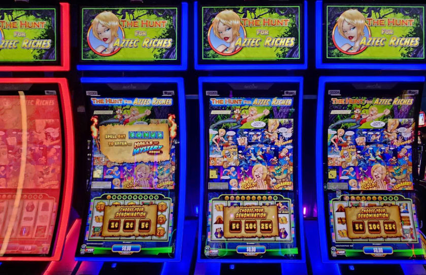Three slot machines with the theme "The Hunt for Aztec Riches" at High Winds Casino. Each machine displays colorful graphics with options to choose the denomination (50, 100, or 250) and a bonus game message to enter "Mystery". The machines are lit with red and blue neon lights.