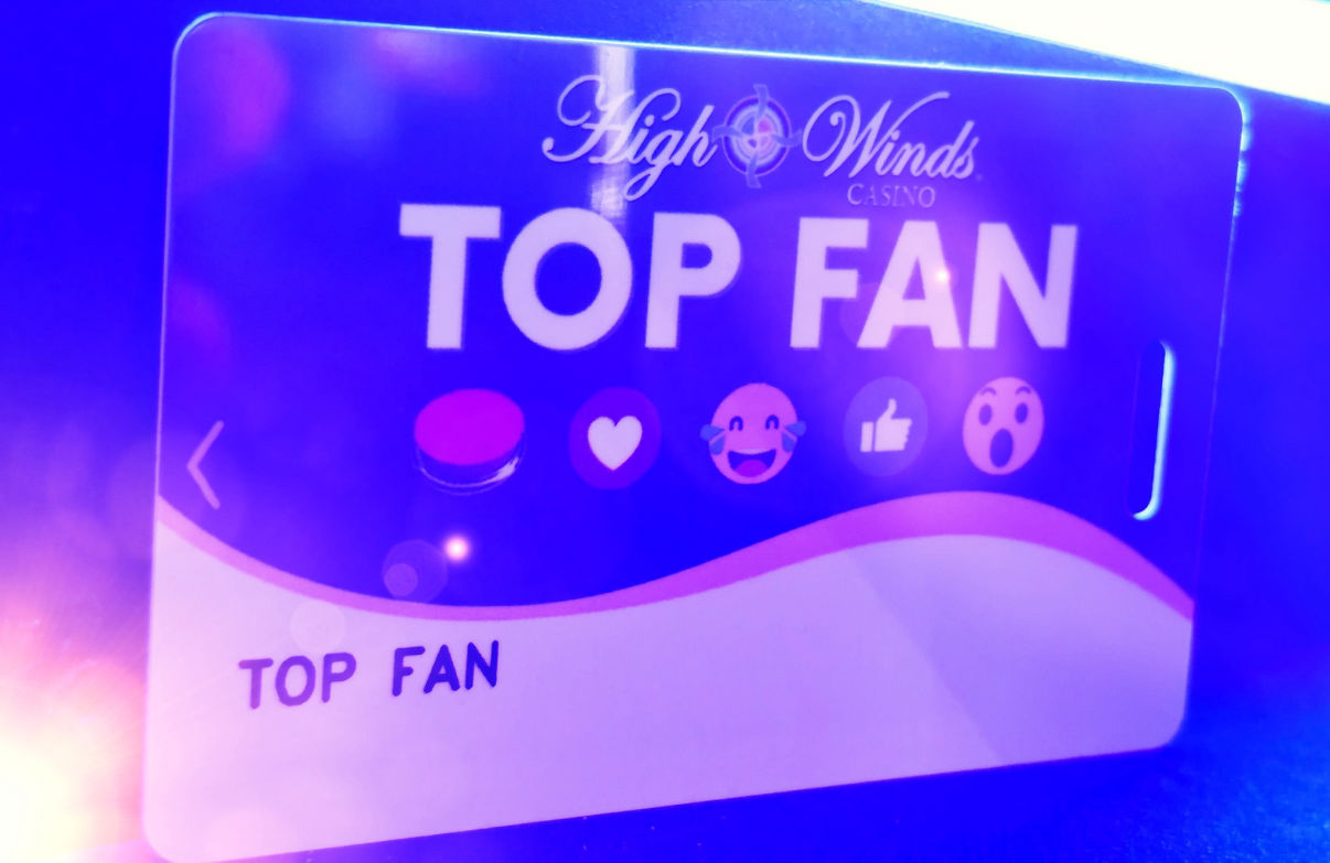 High Winds Casino Top Fan card displayed under vibrant blue and purple lighting. The card features the casino logo, bold 'Top Fan' text, and social media reaction icons, symbolizing exclusive rewards and engagement for top casino supporters."