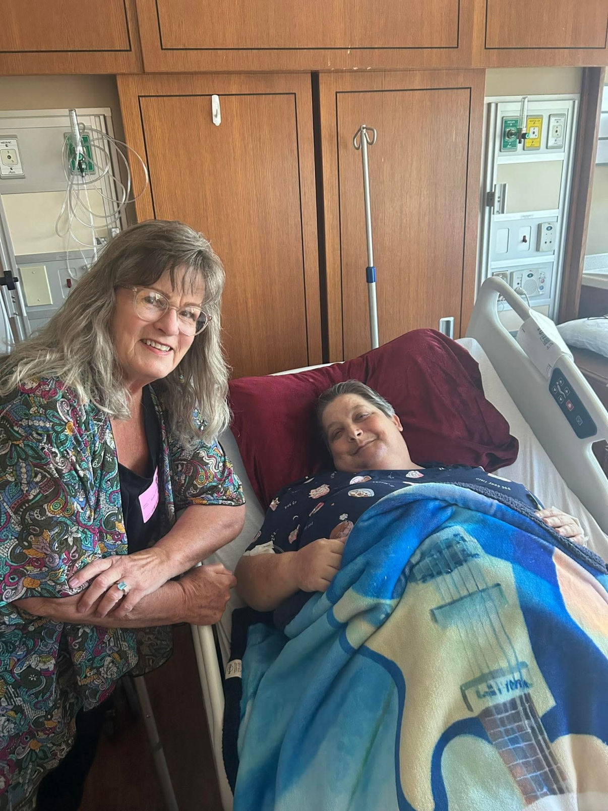 Smiling woman sitting beside a hospital bed with a patient covered by a blue guitar-themed blanket. The patient is resting comfortably, and both appear happy and relaxed in the hospital room setting."