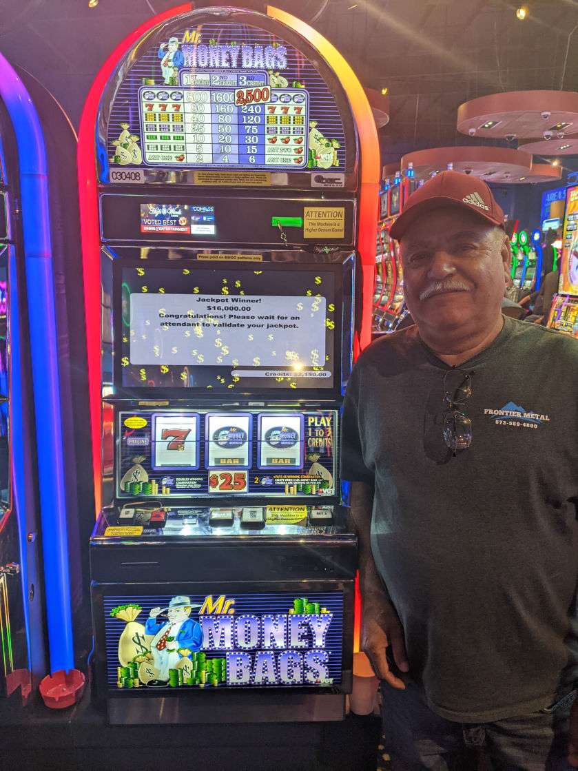 A man stands next to a "Mr. Money Bags" slot machine, displaying a $16,000 jackpot win on the screen. He is smiling while holding a small ticket with the slot machine's credits and a large sign indicating a jackpot win. The vibrant casino setting is visible in the background.